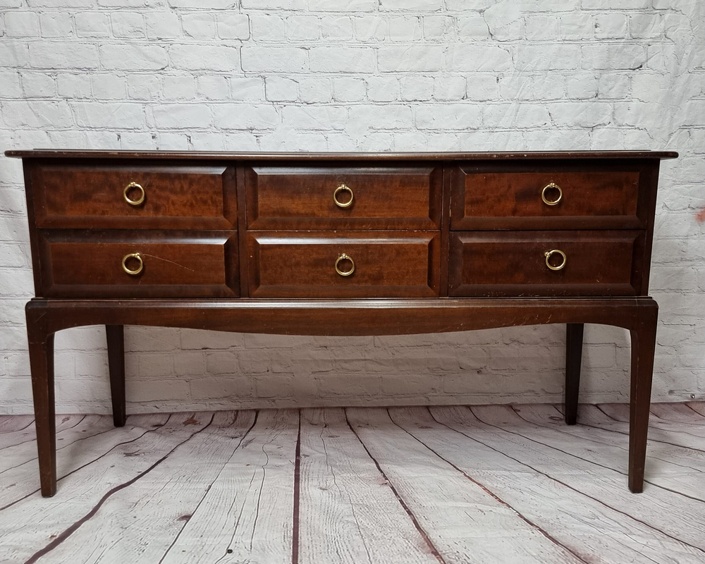 A dark wood sideboard with six drawers, each featuring a gold circular handle, stands against a white brick wall. The sideboard has long, slender legs and a smooth finish, with the wood's grain and texture visible. The floor is made of distressed wood planks.