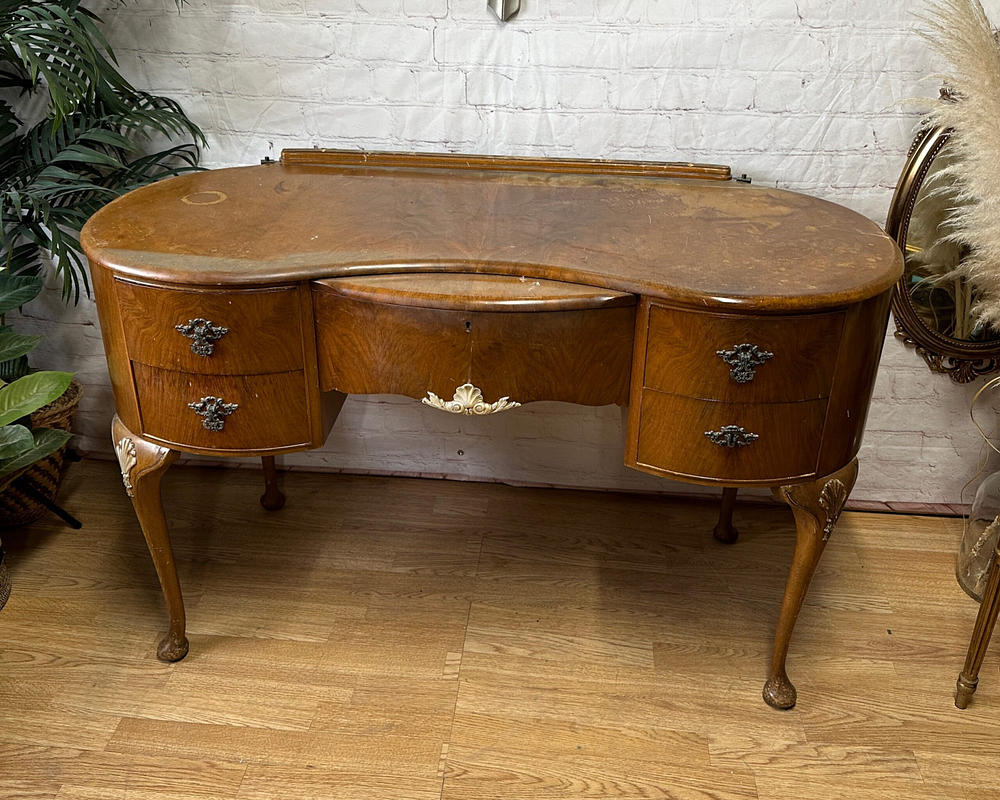 Antique wooden vanity with a curvaceous front, featuring four drawers with intricate metal handles. The vanity has cabriole legs and a light-colored wooden top with visible grain. It stands against a white brick wall, with plants and a decorative mirror nearby.