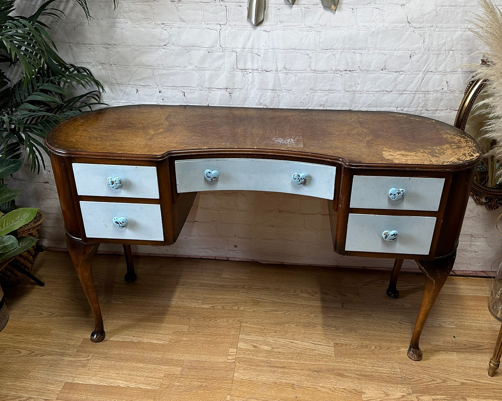 An antique wooden desk with a curved front and six light blue drawers featuring round blue knobs. The desk has cabriole legs and a slightly worn, brown wood finish. It is placed on a wooden floor in front of a white brick wall, surrounded by plants and mirrors.