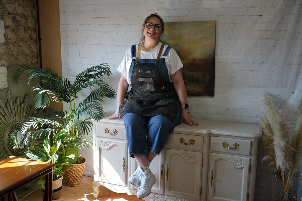 A woman wearing glasses, a white shirt, and denim overalls with paint splatters sits on a white ornate dresser. She is smiling and surrounded by plants. A painting hangs on the brick wall behind her. Sunlight illuminates the scene from the left.