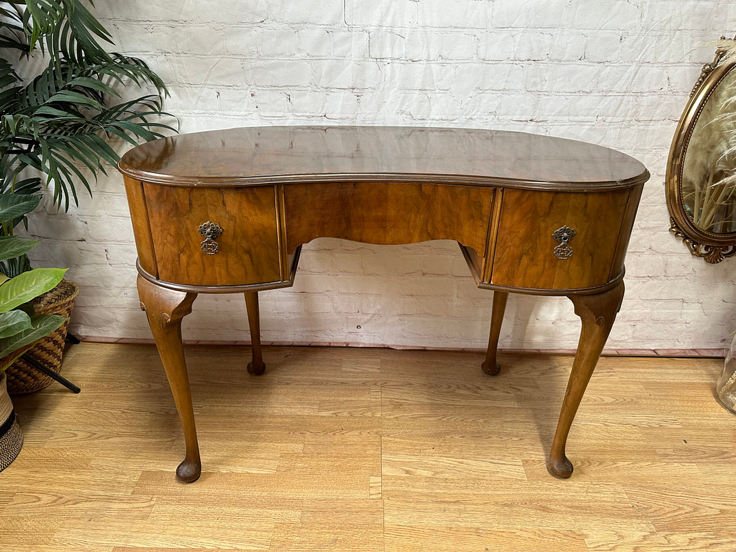 An antique wooden writing desk with curved edges, slender legs, and intricate metal handles stands on a hardwood floor. The background features a white brick wall, a decorative mirror, and green potted plants, creating a classic and elegant setting.