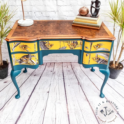 A vintage wooden desk with yellow and teal painted drawers featuring a botanical design. The curved legs and carved details add an elegant touch. On top are stacked books, a lamp, and a decorative sculpture. Potted plants are placed on both sides, and a white brick wall serves as the backdrop.