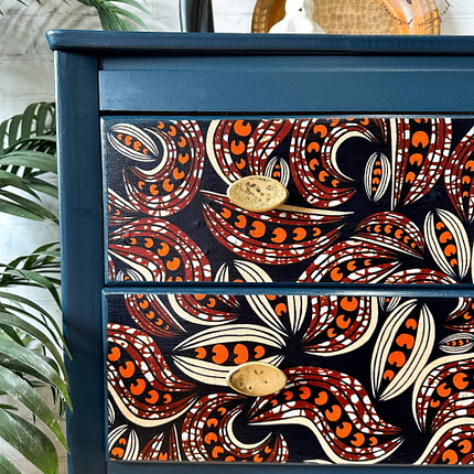 A close-up of a two-drawer cabinet painted in deep blue with vibrant red, orange, and white paisley patterns on the drawer fronts. The drawers have oval-shaped, brass handles. In the background, there are green leaves and a textured wall.