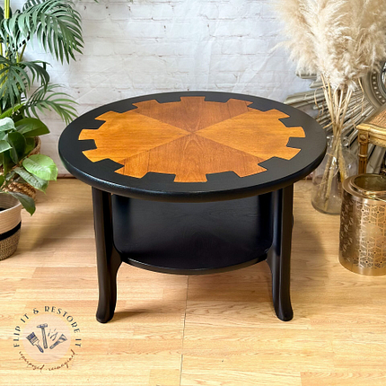 Presenting the MCM Circular Round 'Cog' Coffee Table by Nathan Furniture, a striking piece with a distinctive two-tone design. The center features a natural wood finish with an elegant starburst pattern, while the outer edge is hand-painted in black. This stunning table stands on black legs and complements any decor beautifully when placed on a wooden floor, surrounded by plants and decorative items.