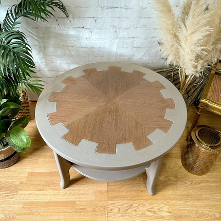 The MCM Circular Round 'Cog' Coffee Table by Nathan Furniture, hand-painted in soft khaki beige, features a unique gear-shaped design in the center with contrasting light and dark wood grains. This wooden side table stands on sturdy legs and is beautifully styled amidst greenery and dried pampas grass against a white brick wall.