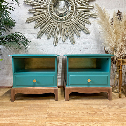 A pair of Stag Minstrel bedside tables with single drawers and open shelves stand side by side on a wooden floor. A decorative mirror with a sunburst design is mounted on the white brick wall behind them. Pampas grass and leafy plants are placed around the furniture.