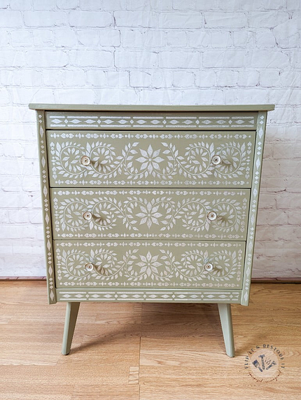 The Faux Bone Inlay Stencil Hand-Painted Vintage Chest of Drawers is a small, green wooden chest with three drawers, each adorned with intricate white floral patterns. The chest stands on four tapered legs and is showcased against a white brick wall background on a hardwood floor.