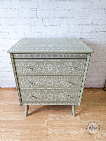 A small Faux Bone Inlay Stencil Hand-Painted Vintage Chest of Drawers in green, featuring three drawers adorned with intricate white floral and geometric patterns. The dresser stands on tapered legs against a white brick wall, with a textured wood floor beneath.