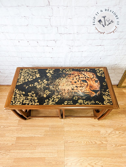 The G Plan Astro Long John Teak Nest Of Tables boasts a bold design on the tabletop, showcasing a leopard's head with a black background adorned with gold spots, creating a striking contrast. The table is elegantly displayed on a light wooden floor against a white brick wall backdrop.