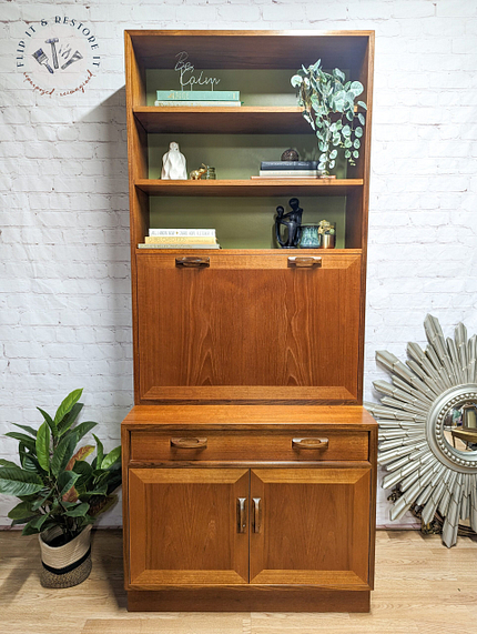 A G-Plan Sierra Tall Teak Bookcase/Drinks Cabinet Mid Century with a cabinet at the bottom and shelves at the top, adorned with various decorative items like books, potted plants, and small statues. A sunburst mirror and a large potted plant sit to the right and left of the cabinet, respectively.