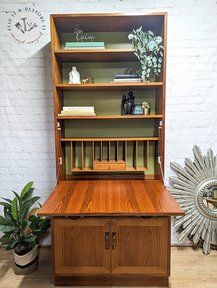 A G-Plan Sierra Tall Teak Bookcase/Drinks Cabinet Mid Century stands against a white brick wall. It features shelves adorned with various decorations, greenery, books, and artistic items. The cabinet door is open, showcasing the stylish interior, while plants decorate the floor to the left and a sunburst mirror is positioned to the right.