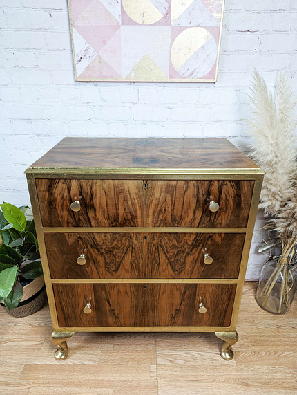 A wooden dresser with three drawers featuring brass handles and edges, placed on a wooden floor. The dresser stands against a white brick wall, adorned with a geometric pattern artwork above. Plants and pampas grass decor surround the furniture.