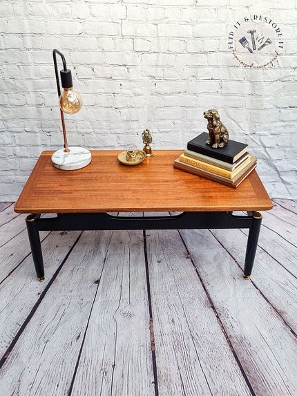 The G-Plan Librenza Mid Century Modern Black and Tola Vintage Coffee Table from the 1950s, adorned with a decorative lamp featuring a unique light bulb, gold accents, and a marble base, sits against a white brick wall with a circular logo. On the table are two gold sculptures, a small tray, and a stack of books.