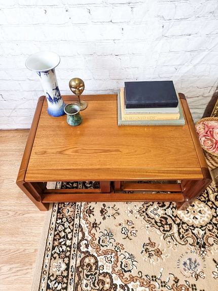 The Mid Century Teak Nest of Tables, consisting of a coffee table and two side tables, is decorated with a blue and white vase, a small brass globe candle holder, a green ceramic cup, and a stack of books. The set is arranged on a patterned carpet against a white brick wall. Pink flowers rest on the floor beside the tables.