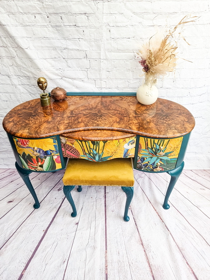 The Queen Anne Painted Burr Walnut Bedroom Set comprises a vintage wooden vanity table with curved edges and a marble-like top. The front showcases vibrant painted plant motifs, and an upholstered yellow velvet stool is nestled underneath. The tabletop is adorned with various decorative items, including a vase filled with dried flowers.