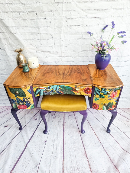 The Queen Anne Painted Walnut Bedroom Set, featuring a dressing table/vanity and stool, stands against a white brick wall and light wooden floor. The vanity boasts colorful, tropical-themed drawer fronts complemented by purple-painted legs. Atop the vanity are candles, a small pineapple decoration, and a purple vase holding an arrangement of flowers.