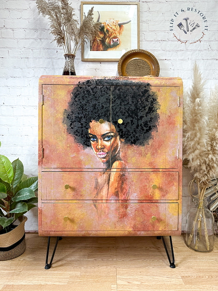 A hand-painted Tallboy cabinet features a vibrant painting of a woman with curly hair against a warm-toned background. The cabinet rests on black hairpin legs and is surrounded by various plants, a decorative plate, and a framed picture of a cow on a white brick wall.