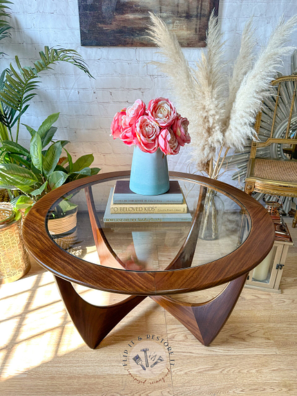 A G-Plan Astro Round Coffee Table Mid-Century Vintage with a wooden base stands in a well-lit room. On top of the table is a blue vase filled with pink flowers and two books. Surrounding the table are potted plants, a wicker chair, and decorative pampas grass.