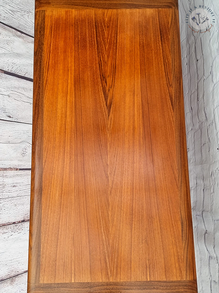 A G Plan Astro Mid Century Modern Vintage Teak Coffee Table from the 1960s, showcasing a smooth, polished surface with a rich, warm brown hue and distinct wood grain patterns. The table is set against a white-washed wooden background that features visible textures and a decorative emblem in the top right corner.