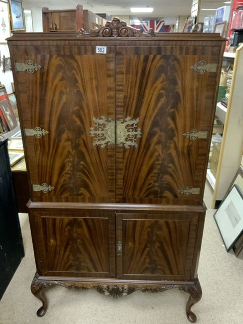 A tall, antique wooden cabinet with ornate metal accents and detailed carving at the base is displayed in a room filled with various other pieces of furniture and framed artwork. The cabinet features two large doors on the upper section and two smaller doors on the lower section.