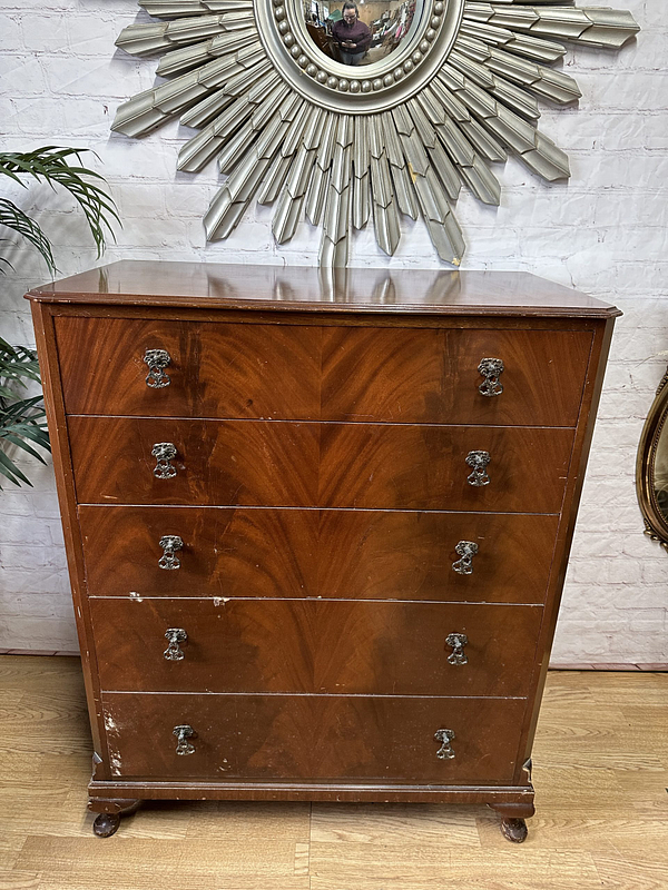 A wooden chest of drawers stands against a white brick wall. It has five drawers with vintage metal handles. Above it hangs a round mirror adorned with a sunburst frame. A green plant is partially visible on the left, and a circular mirror in a gold frame on the right.