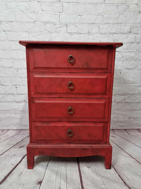 A small red wooden dresser with four drawers and brass ring handles stands on a wooden floor against a white brick wall background.