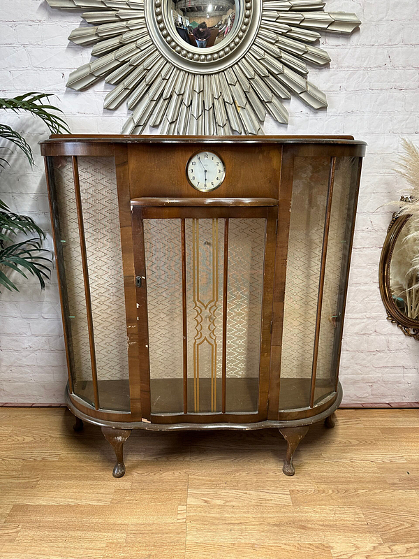 A vintage wooden display cabinet with curved glass sides stands on cabriole legs. It features a clock at the center top and ornate patterned detailing on the front glass panels. Behind it is a brick wall with a decorative round mirror and nearby plants.