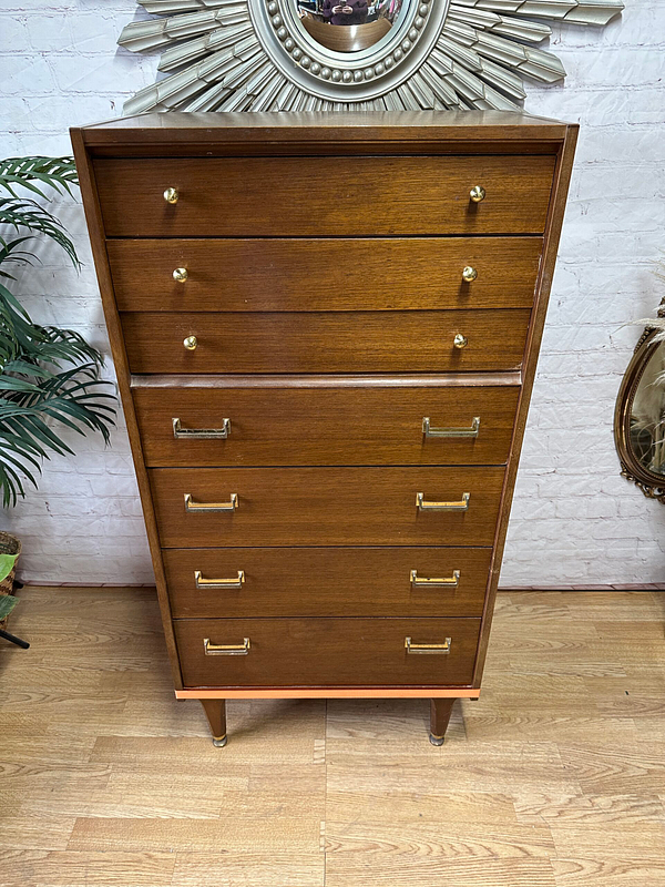 A tall wooden dresser stands against a white brick wall. The dresser has seven drawers, the top three smaller with round knobs and the bottom four larger with rectangular handles. A decorative framed mirror and a green plant are on either side.