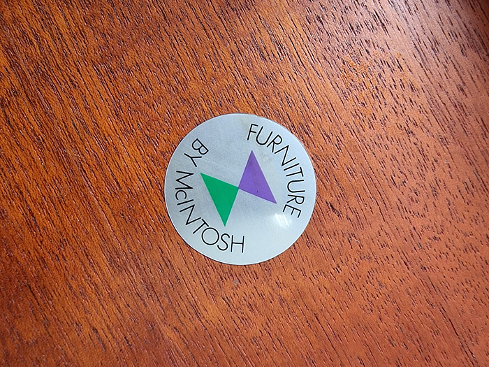 A silver circular sticker with green and purple overlapping triangles in the center. The text around the edge reads "Furniture by McIntosh." The sticker is attached to an Ercol Saville Ash Long Coffee Table with Smoked Glass Mid Century Vintage, featuring a rich, brown finish.