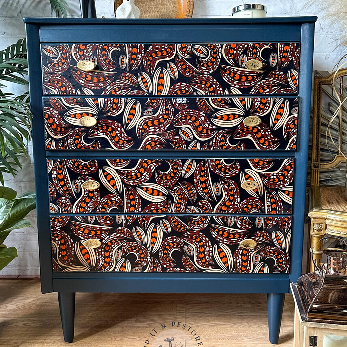 A vibrant navy blue chest of drawers with an intricate, colorful paisley design on the drawer fronts in shades of red, orange, and white. The surroundings include plants and various decor items, creating a stylish and lively atmosphere.