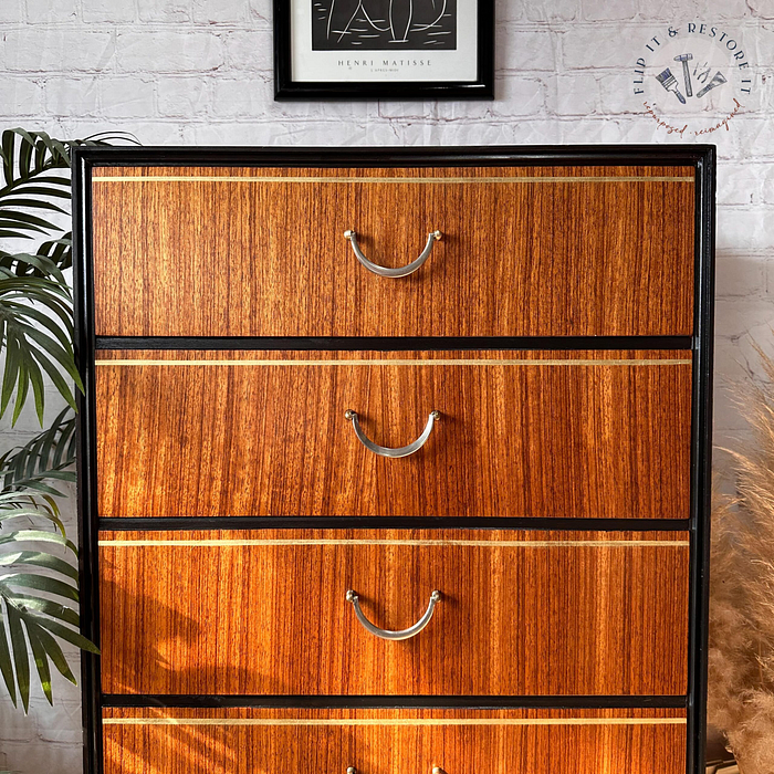 An Exquisite Mid Century MCM Walnut Tallboy with 6 drawers by Meredew, featuring a black frame and metal handles. The chest boasts drawers that showcase a warm, rich wood grain pattern. Above it, a framed abstract art print adorns the brick wall, while a tall houseplant is partially visible on the left side.