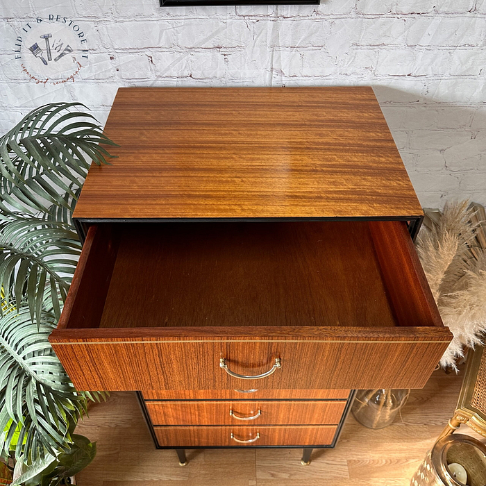 The Exquisite Mid Century MCM Walnut Tallboy 6 Drawers Chest Of Drawers by Meredew is seen with one drawer open. The chest has a polished finish and three visible drawers with golden handles. Surrounding the chest are a green potted plant on the left, a wicker chair on the right, and a white brick wall behind.