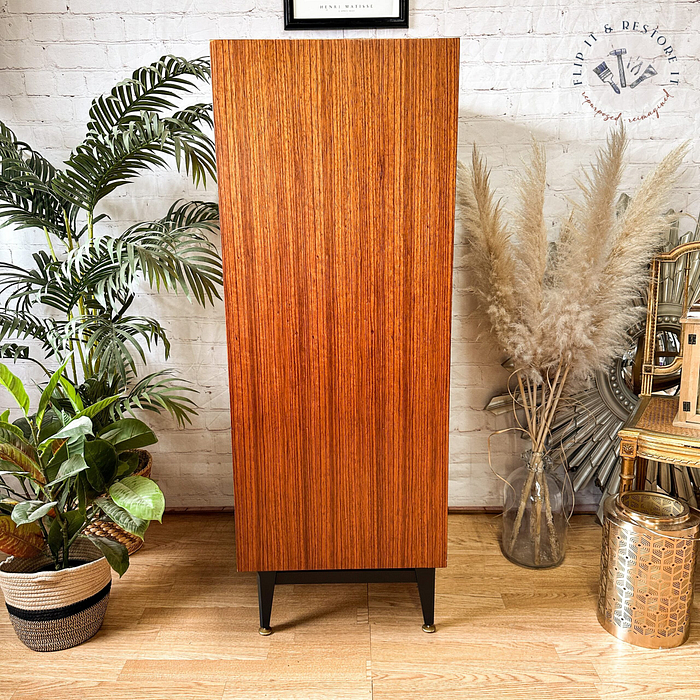 The Exquisite Mid Century MCM Walnut Tallboy 6 Drawers Chest Of Drawers by Meredew stands on black legs against a white brick wall. It is flanked by green plants on the left and dried pampas grass in a vase on the right. A small, mirrored stool is partially visible on the right side.