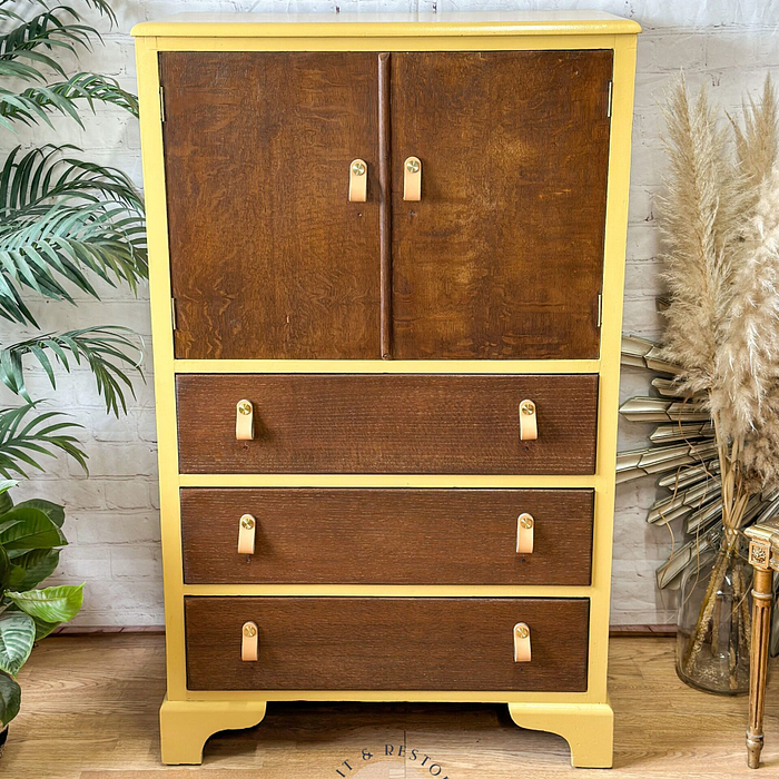 The Hand-painted Tallboy Linen Cupboard, featuring a mid-century modern design, showcases a striking yellow frame complemented by dark wood drawers. This upcycled cabinet includes three spacious drawers with elegant leather handles and two small upper doors. Positioned on a hardwood floor, it is flanked by lush plants like palm and pampas grass, making it ideal for bedroom or kitchen storage.