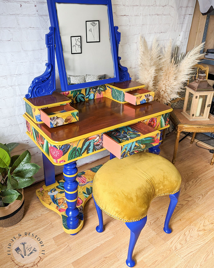 The Vintage Victorian Mahogany Duchess Dressing Table Painted Bedroom Set includes a beautifully painted vanity in blue and yellow with floral patterns, featuring several small drawers. Accompanied by an upholstered stool with a yellow velvet seat, the set is elegantly positioned on a wooden floor, surrounded by potted plants and decor.