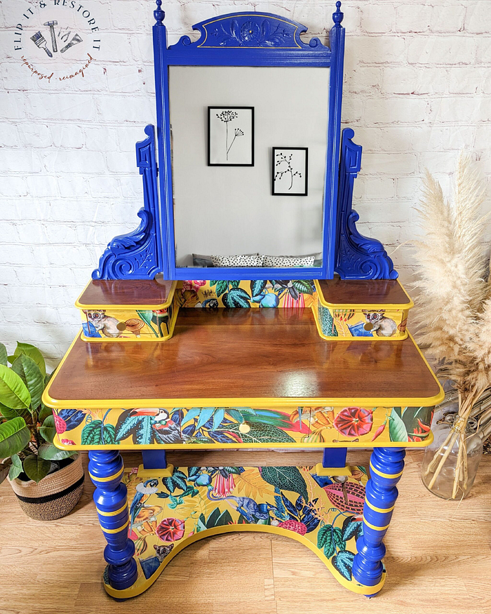 A vibrantly painted Vintage Victorian Mahogany Duchess Dressing Table with a large mirror. The mirror's frame and legs are primarily blue with intricate carvings, while the drawers and lower shelf feature bold floral designs in yellow, green, and red. Various plants and framed art are in the background.