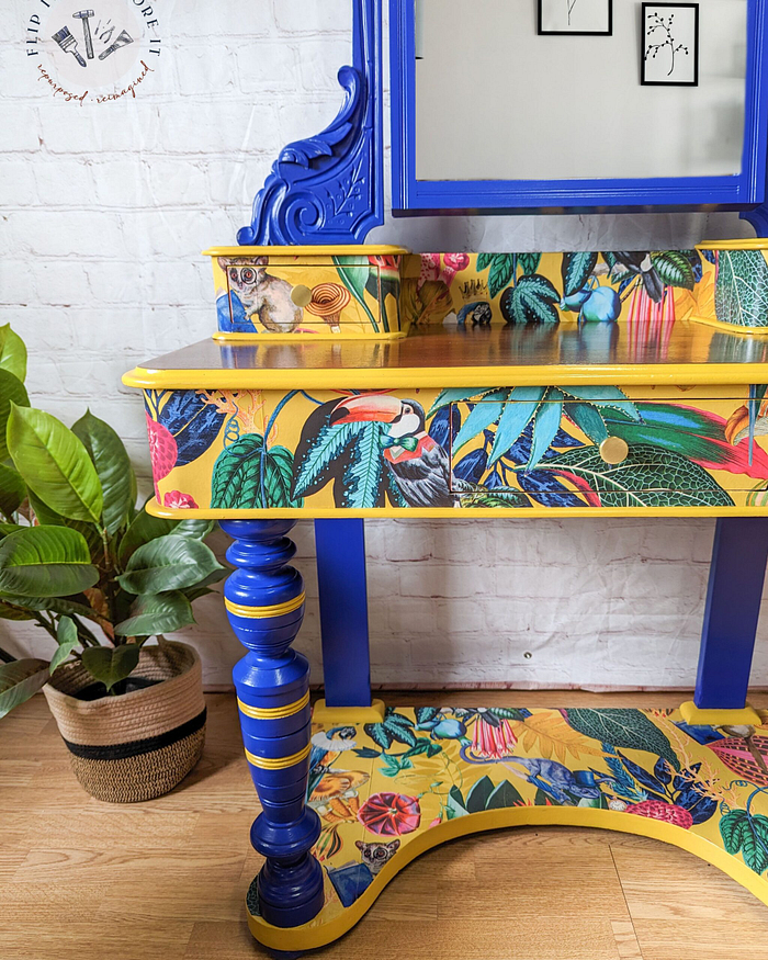 A vibrant, ornately decorated Vanity from the Vintage Victorian Mahogany Duchess Dressing Table Painted Bedroom Set sits against a white brick wall. The blue and yellow surfaces of the dressing table feature tropical-themed illustrations of toucans, foliage, monkeys, and various colorful plants. A potted plant rests on the floor to the left of this charming vanity.