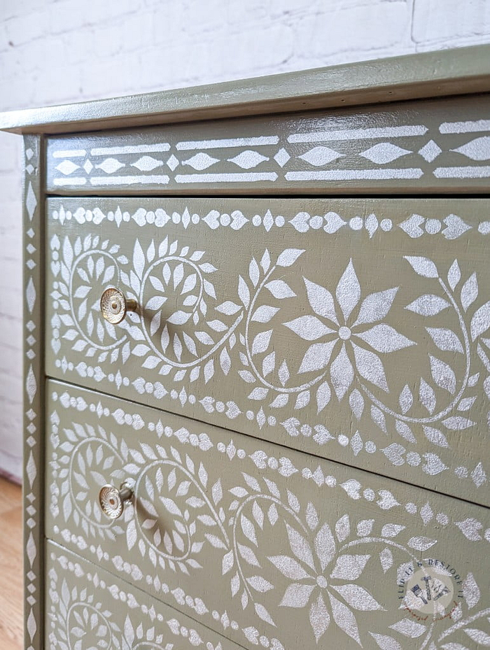 A Faux Bone Inlay Stencil Hand-Painted Vintage Chest of Drawers in green, adorned with intricate white floral and geometric patterns, featuring three drawers with small, round metallic handles. The background showcases a white brick wall, and part of a wooden floor is visible at the bottom.