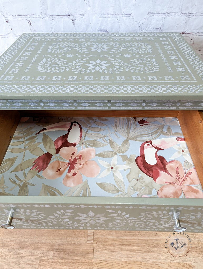 A wooden drawer from the Faux Bone Inlay Stencil Hand-Painted Vintage Chest of Drawers is partially open, showcasing an intricate floral and bird pattern inside. The exterior of the drawer features a decorative green and white design with geometric and floral elements. The drawer is placed on a wooden floor against a white brick wall background.