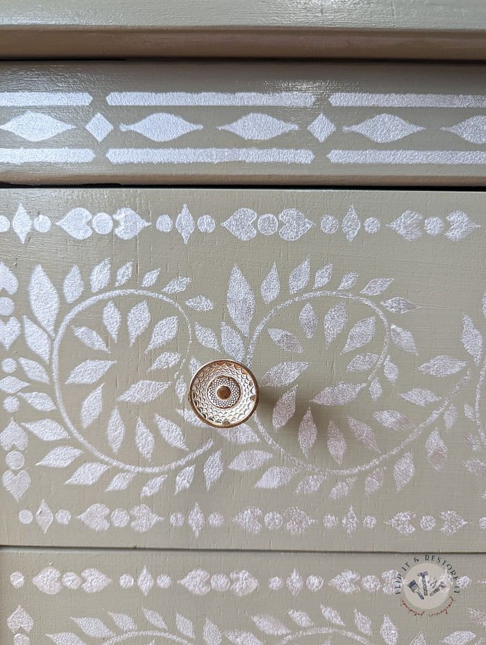 A close-up image of a drawer from the Faux Bone Inlay Stencil Hand-Painted Vintage Chest of Drawers, featuring intricate white floral and leaf patterns on a beige background. The drawer has a single, circular, bronze-colored knob in the center. A small logo is visible in the bottom right corner of the image.