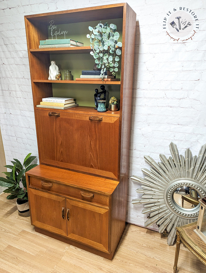 The G-Plan Sierra Tall Teak Bookcase/Drinks Cabinet Mid Century showcases a variety of items with its two lower cabinets and multiple shelves, displaying books, a small statue, a decorative candle, and potted plants. Set against a white brick wall background, the scene is enhanced by the presence of a sunburst mirror on the right.