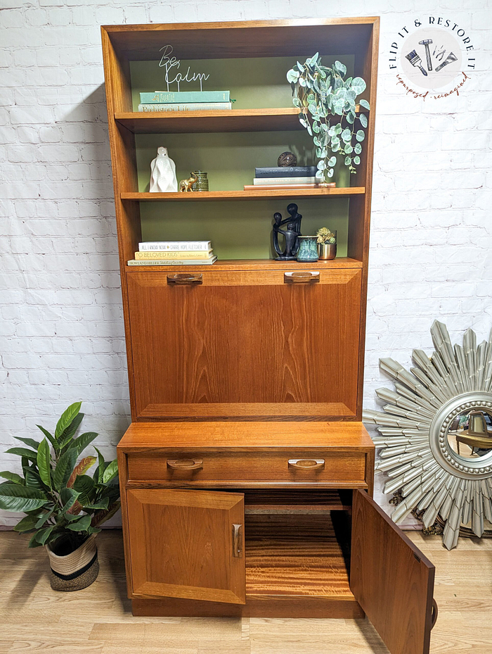 The G-Plan Sierra Tall Teak Bookcase/Drinks Cabinet Mid Century features open upper shelves displaying decor items such as books, a potted plant, a statue, and a "Be Calm" sign. Below is a fold-down desk workspace and a storage compartment with one door ajar. Positioned to the right on the floor is an elegant decorative mirror.