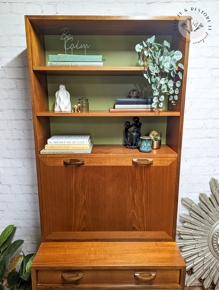 A G-Plan Sierra Tall Teak Bookcase/Drinks Cabinet Mid Century against a white brick wall features open shelves and a closed cabinet at the bottom. The top shelf holds a "Be Calm" sign, book, and potted plant. The middle shelf displays books, a small sculpture, and another plant. Greenery is also visible on the sides of the cabinet.