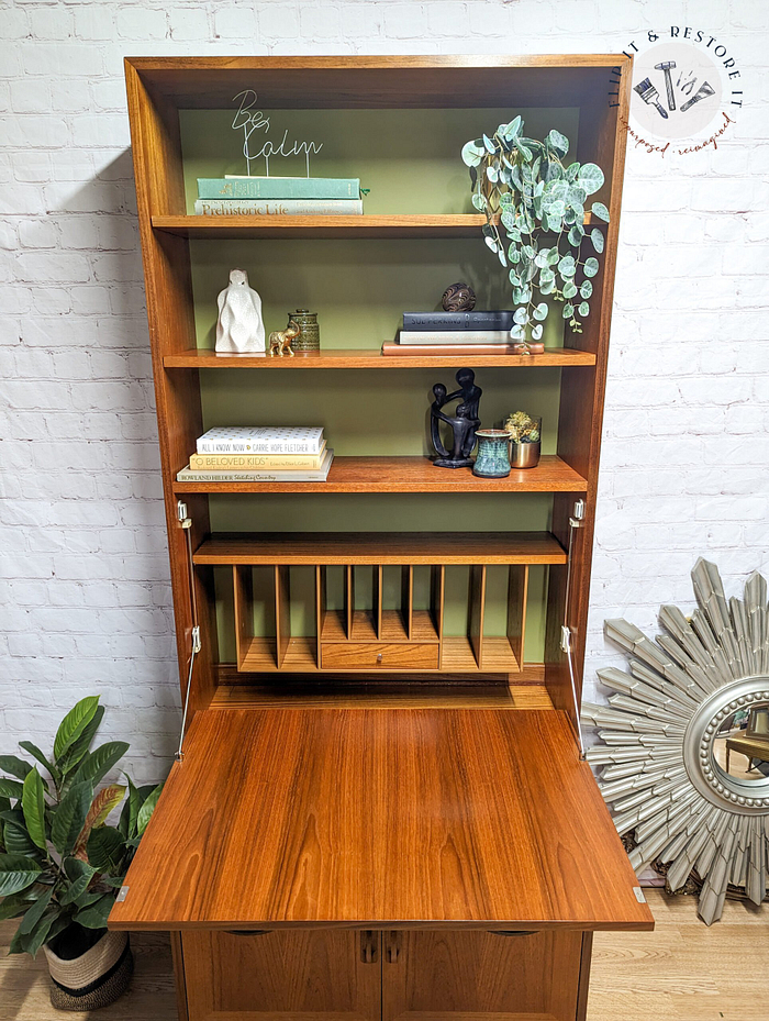 A G-Plan Sierra Tall Teak Bookcase/Drinks Cabinet Mid Century with an open top displaying decorative items such as books, a small vase, a statue, and plants. The bottom section includes vertical slots for organizing papers. The bookcase is surrounded by a decorative mirror, a potted plant, and set against a brick wall background.