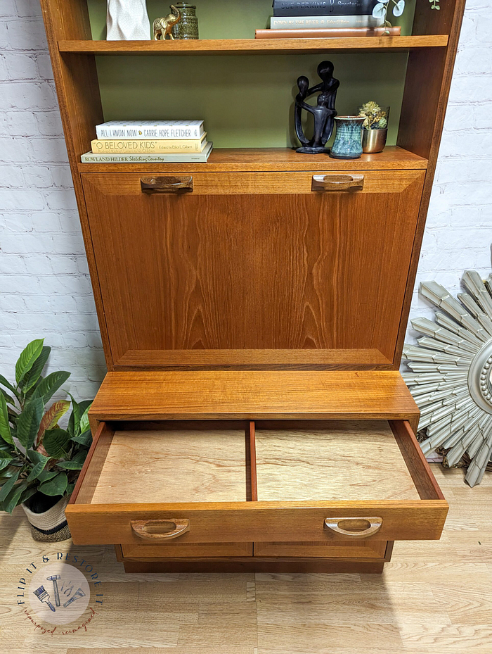 A G-Plan Sierra Tall Teak Bookcase/Drinks Cabinet Mid Century stands against a white brick wall. The top shelf holds decorative items and books, while the middle section features a drop-down door revealing two compartments. The bottom section consists of two closed drawers. A plant is positioned on the left and a sunburst-shaped mirror adorns the right side.