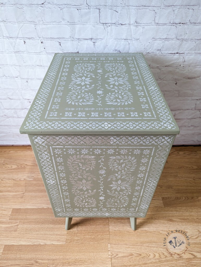 A light green Faux Bone Inlay Stencil Hand-Painted Vintage Chest of Drawers adorned with intricate white floral and geometric patterns on the top and front. The chest stands gracefully on four legs, placed on a wooden floor against a white brick wall, with the "Lily & Design" logo visible in the bottom right corner.