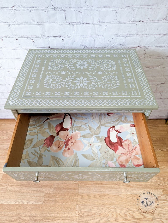 A Faux Bone Inlay Stencil Hand-Painted Vintage Chest of Drawers, featuring a square green design with a patterned white floral motif on the top and sides. This chest boasts an open drawer adorned with decorative paper showcasing birds and floral designs. The chest is displayed against a white brick wall on a wooden floor.