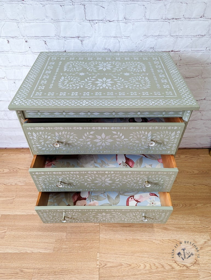 A small Faux Bone Inlay Stencil Hand-Painted Vintage Chest of Drawers stands against a white brick wall. The chest features intricate white stencil designs on the top and drawers. Two lower drawers are pulled open, revealing a floral-patterned lining inside. The wood floor complements the chest.