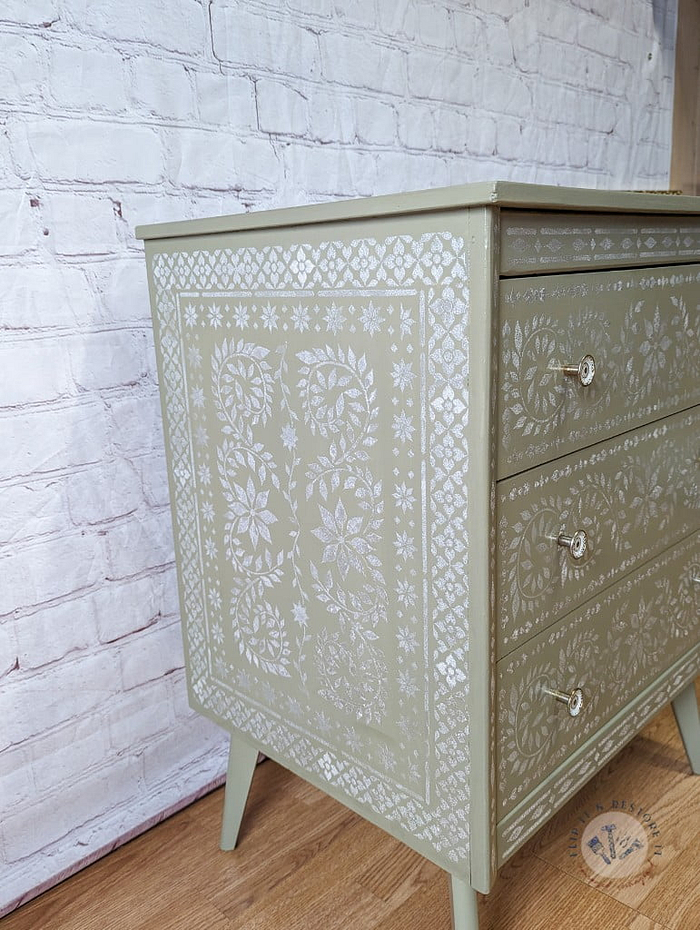 A Faux Bone Inlay Stencil Hand-Painted Vintage Chest of Drawers, showcasing intricate white floral and geometric designs against a light green backdrop, stands gracefully against a white brick wall. The chest features three drawers, each adorned with a small round metallic knob. The wooden floor beneath enhances the warmth of the setup.