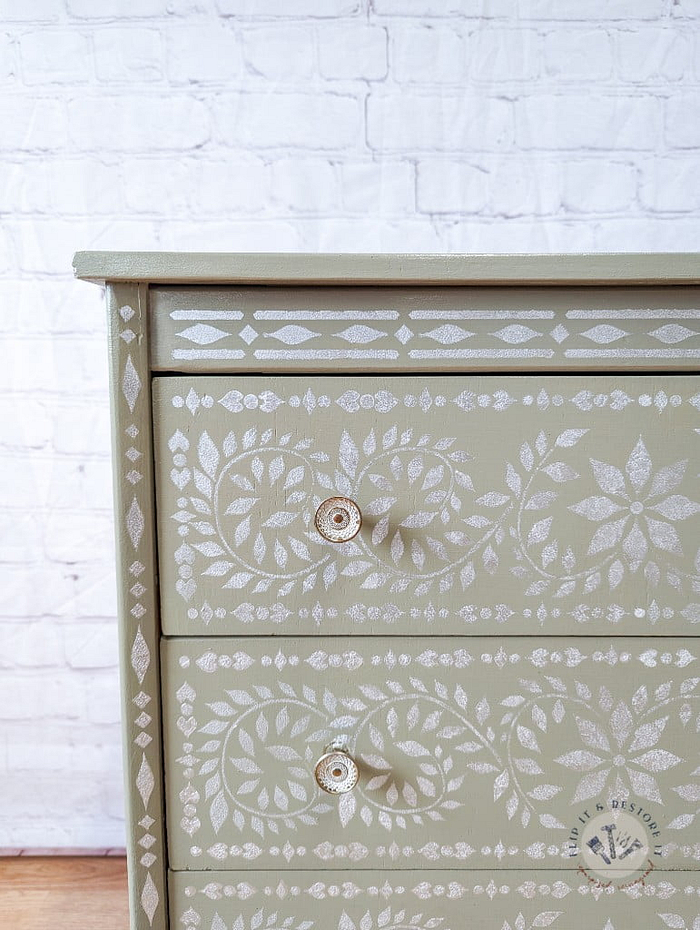 The Faux Bone Inlay Stencil Hand-Painted Vintage Chest of Drawers is a light green dresser featuring three drawers, each adorned with intricate white floral patterns and round white knobs. Set against a white brick wall, the muted color and detailed design of the furniture create a striking contrast.